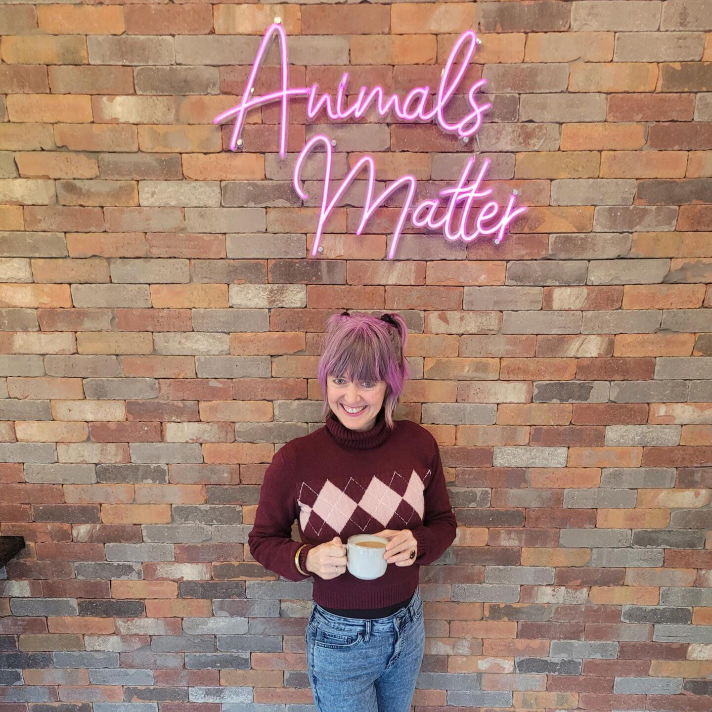 Animals Matter Sadie Freely holding a cup of coffee with neon pink Animal Matters sign above her on brick wall